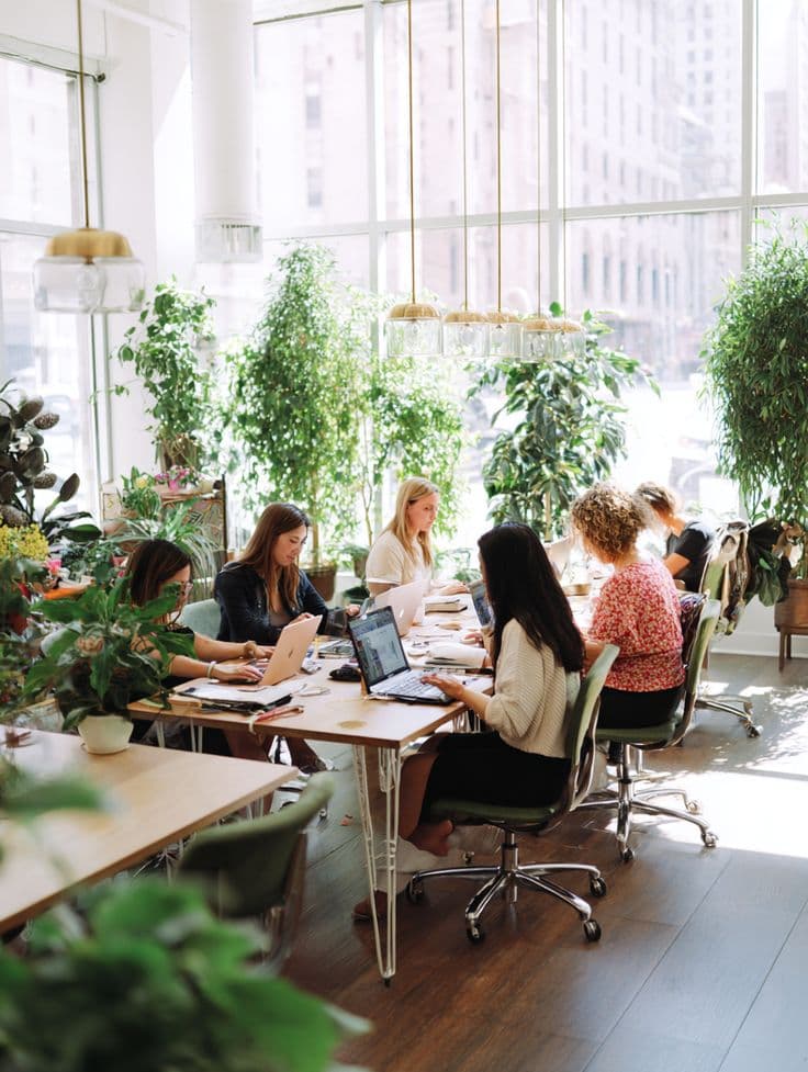 People working at a long shared table in a bright, plant-filled coworking space